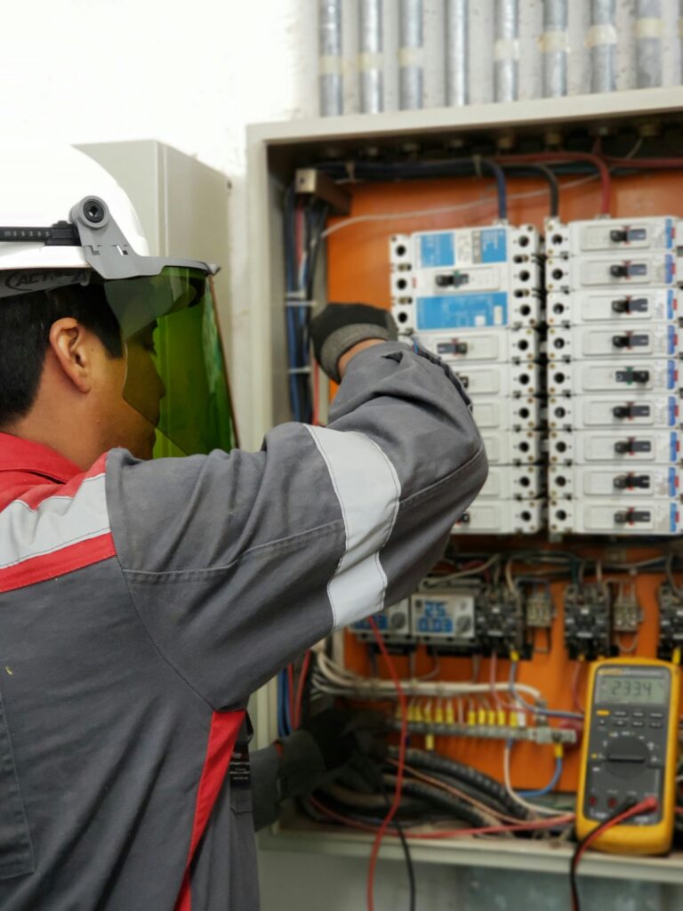 An electrician wearing a white hard hat and an arc flash face shield inspects a large industrial electrical panel with rows of circuit breakers.