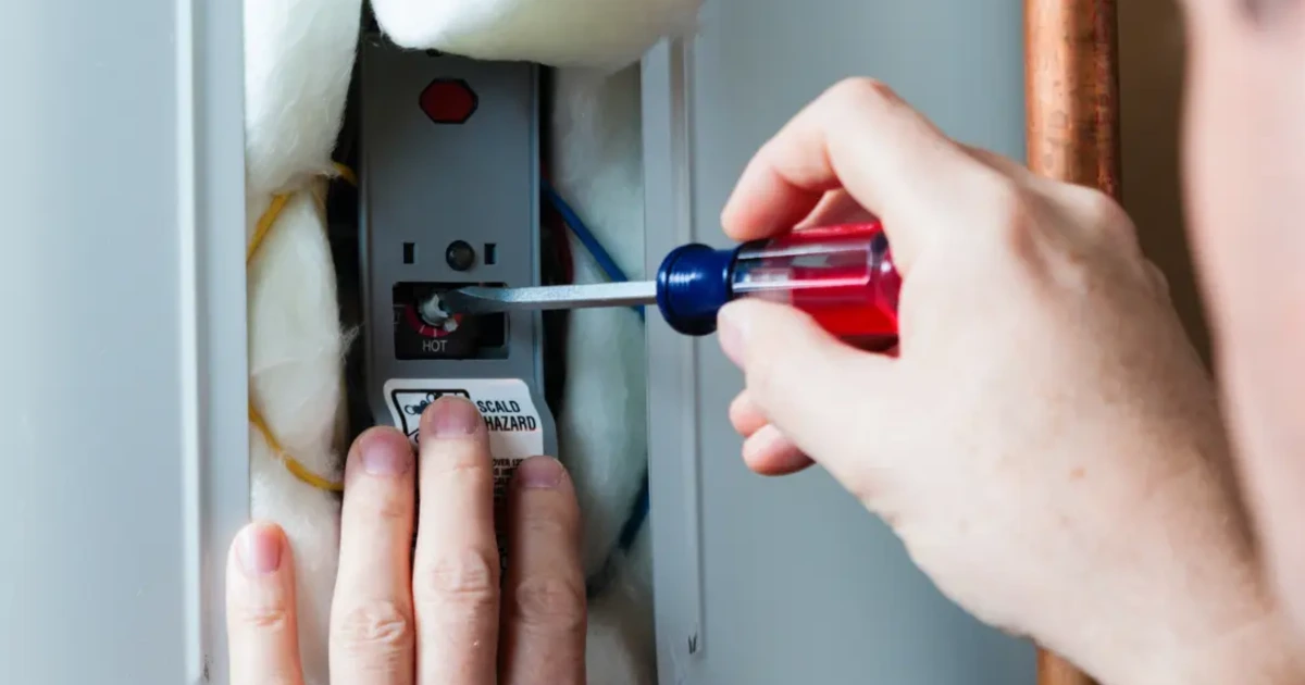 Technician adjusting the thermostat inside an electric storage tank water heater with the access panel open and the scald hazard label visible